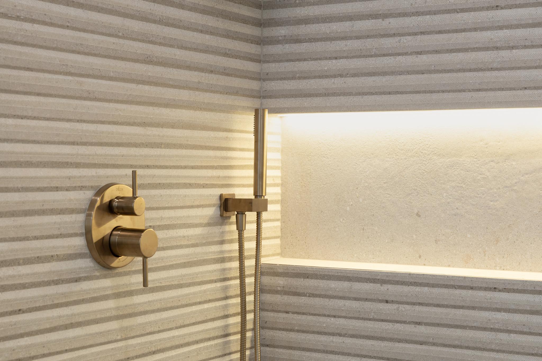 Close-up of premium brushed bronze shower fixtures and an illuminated custom shower niche against bespoke fluted stone tile in a luxury San Jose custom home by Ritz Builders.