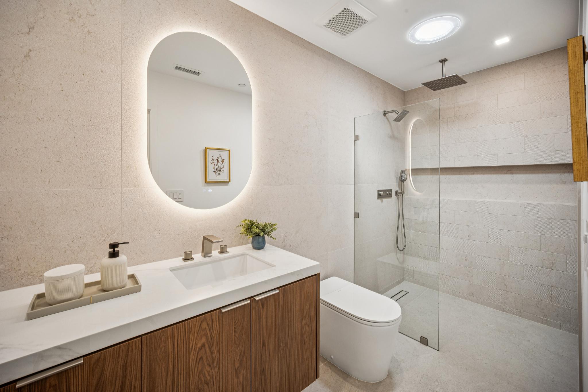 Contemporary luxury guest bathroom in a San Jose custom home, highlighting a floating wood vanity, an illuminated oval mirror, and a zero-threshold walk-in shower beneath a custom skylight by Ritz Builders.