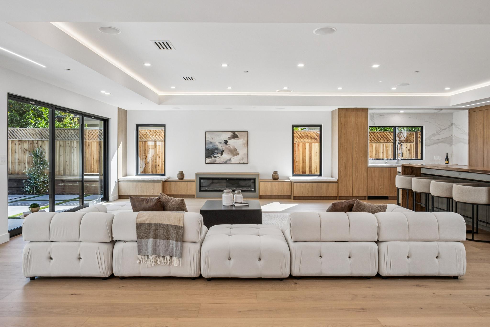 Open-concept modern living room in a Willow Glen luxury home, featuring a low-profile white modular sectional sofa, custom oak cabinetry built-ins, and a minimalist linear fireplace.