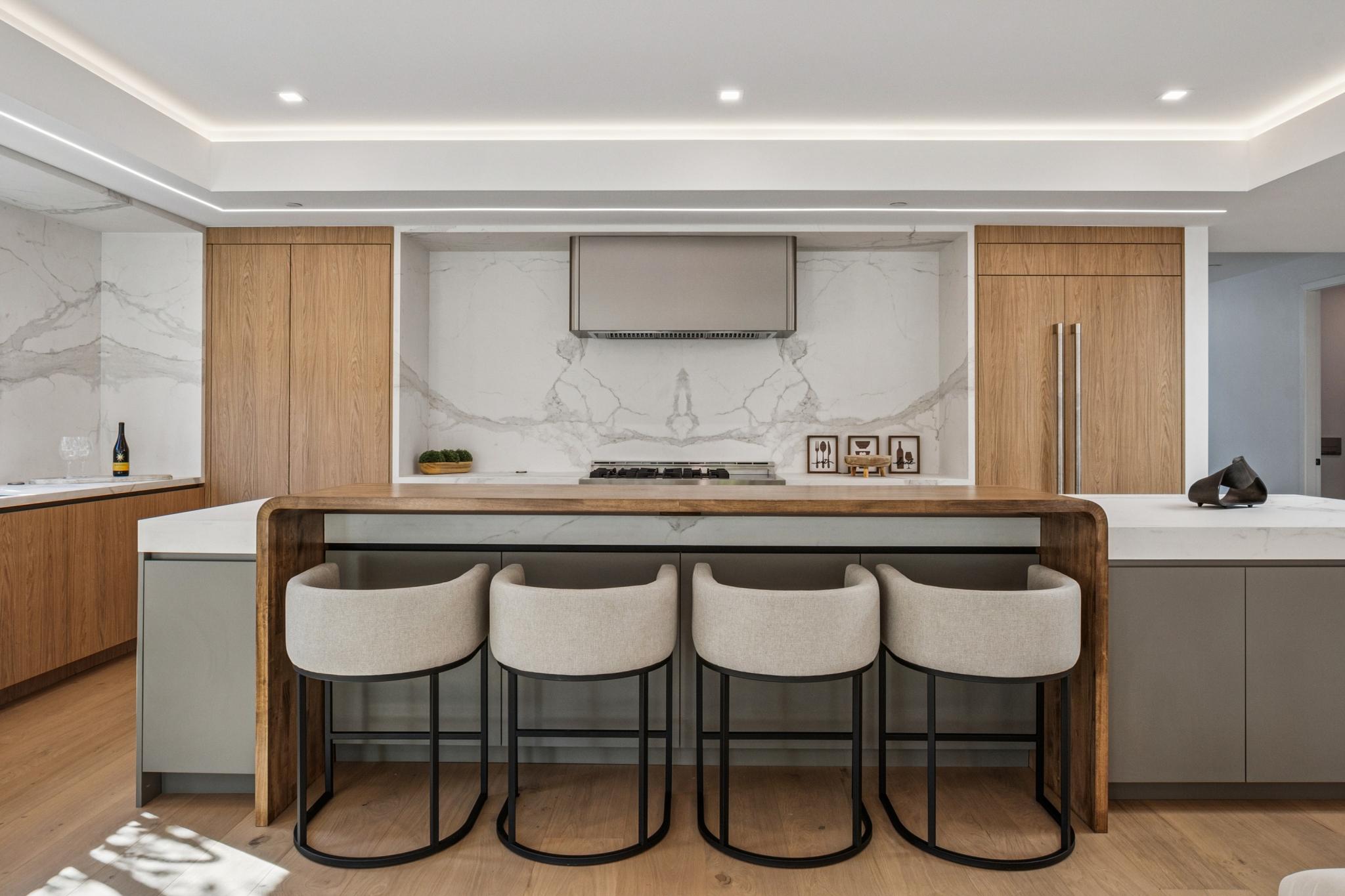 Luxury modern kitchen design in San Jose, featuring a marble waterfall island, custom white oak wood cabinets, integrated appliances, and a unique continuous wood breakfast bar.